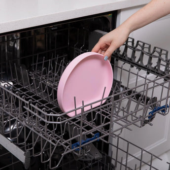 A hand places a Bumkins Silicone Grip Plate: Pink into the top rack of an open dishwasher.