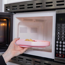 Load image into Gallery viewer, A hand places a Bumkins Silicone Grip Plate: Pink filled with food into an open microwave.
