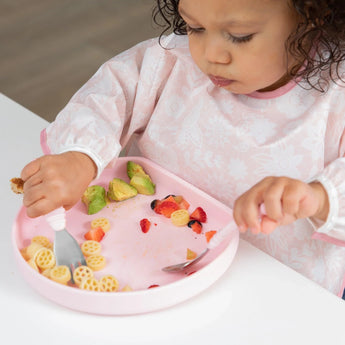 A toddler enjoys pasta, avocado, and strawberries with a fork & spoon from a Bumkins Silicone Grip Plate: Pink on a white table.