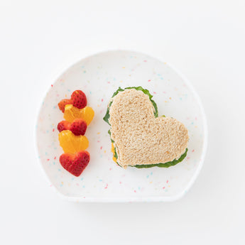 Bumkins Silicone Grip Plate: Vanilla Sprinkle holds a heart-shaped sandwich and heart-shaped pieces of fruit on a white background.
