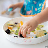 Close-up of a child reaching for fruit from a Bumkins Silicone Grip Plate: Vanilla Sprinkle while wearing a colorful bib.