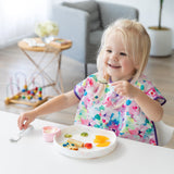 A young girl enjoys food from a Bumkins Silicone Grip Plate: Vanilla Sprinkle with matching utensils, with a toy and chair in the background.