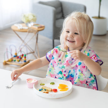 A young girl enjoys food from a Bumkins Silicone Grip Plate: Vanilla Sprinkle with matching utensils, with a toy and chair in the background.