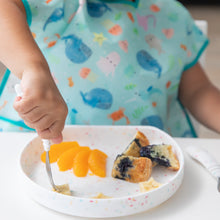 Load image into Gallery viewer, A child's hand uses a fork to eat food from a Bumkins Silicone Grip Plate: Vanilla Sprinkle, wearing a sea creature bib at a white table.
