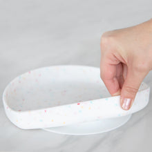 Load image into Gallery viewer, A hand holds a Bumkins Silicone Grip Plate: Vanilla Sprinkle, demonstrating the suction cup bottom on a marble surface.
