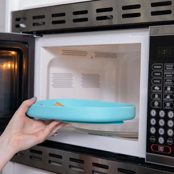 A hand places a Bumkins Silicone Grip Tray: Blue filled with food into an open microwave.