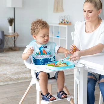A toddler wearing a bib in a high chair enjoys noodles from a Bumkins Silicone Grip Tray: Blue, as a woman nearby holds a fork of food.