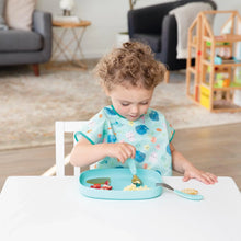 Load image into Gallery viewer, A toddler with curly hair eats from a Bumkins Silicone Grip Tray: Blue at a white table, with toys and a sofa in the background.
