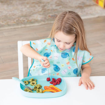 A child in a blue ocean-themed bib enjoys raspberries, pasta, and peppers from a Bumkins Silicone Grip Tray: Blue at a white table.
