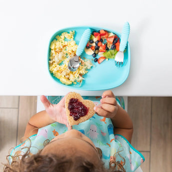 Top-down view of a child eating toast with jam while a Bumkins Silicone Grip Tray: Blue with scrambled eggs and fruit sits on a white table.
