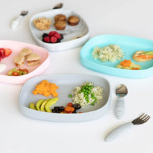 Load image into Gallery viewer, Four plates, including Bumkins Silicone Grip Tray: Gray, are arranged with various foods and utensils on a white background.
