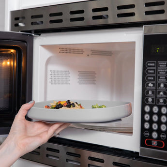 A hand places a Bumkins Silicone Grip Tray: Gray filled with food into an open microwave.
