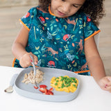 A child in a colorful bib enjoys chicken, strawberries, and macaroni with peas from a Bumkins Silicone Grip Tray: Gray at a white table.