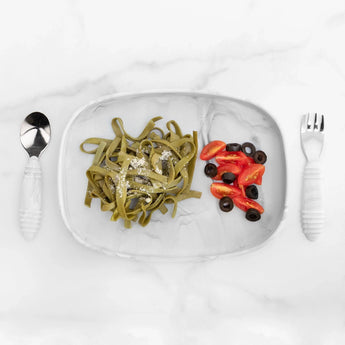 Bumkins Silicone Grip Tray: Marble holds spinach pasta, cherry tomatoes, and olives with matching utensils nearby on a marble background.
