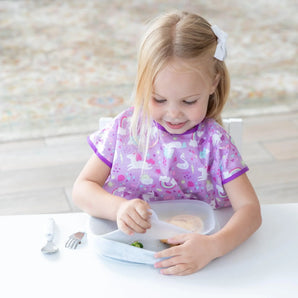 A young blonde child in a purple bib sits at a white table, removing a lid from a Bumkins Silicone Grip Tray: Marble.