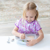 A young blonde child in a purple bib sits at a white table, removing a lid from a Bumkins Silicone Grip Tray: Marble.