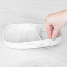 Load image into Gallery viewer, A hand holds a Bumkins Silicone Grip Tray: Marble, demonstrating the suction cup bottom on a marble surface.
