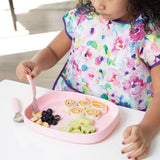 A child with a colorful bib sits at a white table, eating rolled sandwiches and fruit from a Bumkins Silicone Grip Tray: Pink with a fork.