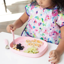Load image into Gallery viewer, A child with a colorful bib sits at a white table, eating rolled sandwiches and fruit from a Bumkins Silicone Grip Tray: Pink with a fork.
