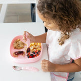 A child in a pink bib eats fruit, toast, and eggs from a Bumkins Silicone Grip Tray: Pink with pink utensils at a white table.
