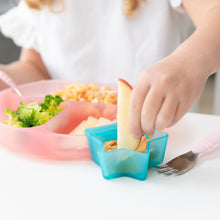 Load image into Gallery viewer, A child dips an apple slice into peanut butter from a Bumkins Silicone Little Dipper Combo 2 Pack: Aqua Jelly at a white table.
