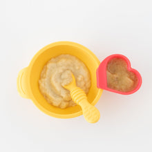 Load image into Gallery viewer, Top-down view of Bumkins Silicone First Feeding Set: Pineapple and a red heart-shaped cup filled with mashed food on a white background.
