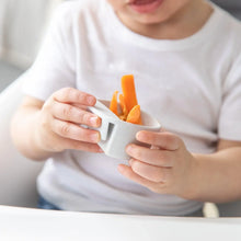 Load image into Gallery viewer, A child in a white shirt holds a marble Bumkins Silicone Little Dipper Round 3-Pack: Taffy filled with carrot sticks.

