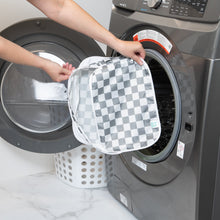 Load image into Gallery viewer, A person holds a Bumkins Packing Cube 2 Pack: Charcoal Check in front of a front-loading washer, with a white laundry basket behind it.
