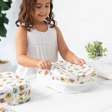 Load image into Gallery viewer, A girl in a white dress smiles, holding a Bumkins Packing Cube 2 Pack: Winnie + Friends, made with waterproof fabric, on the bed.
