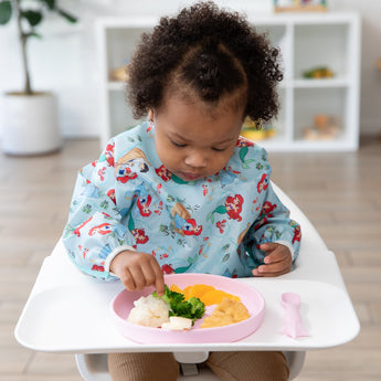A toddler in a bib sits in a high chair, eating from Bumkins Silicone Plate + Spoon Set: Disney Princess with veggies, nuggets, and potatoes.