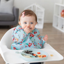 Load image into Gallery viewer, A girl wearing a blue bib smiles as she eats from a Bumkins Silicone Plate + Spoon Set: Disney Princess Ariel in a high chair.
