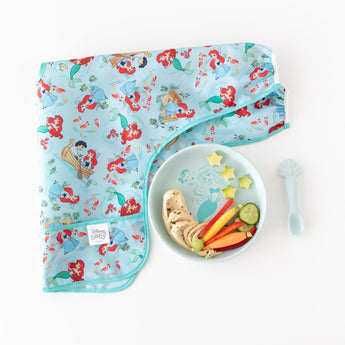 Bumkins Silicone Plate + Spoon Set: Disney Princess Ariel, holding hummus, carrots, and cucumber, sits beside a bib on a white background.