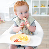A curly red-haired baby in a bib sits in a high chair, eating breakfast from a Bumkins Silicone Plate + Spoon Set: Disney Winnie the Pooh.