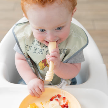 A red-haired baby in a high chair with a bib enjoys bananas and strawberries from Bumkins Silicone Plate + Spoon Set: Disney Winnie the Pooh.