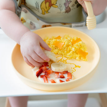 A child wearing a bib in a high chair enjoys breakfast from a Bumkins Silicone Plate + Spoon Set: Disney Winnie the Pooh.