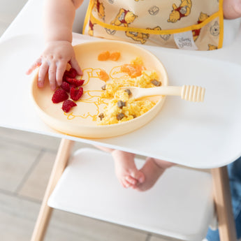 A baby in a high chair reaches for raspberries from Bumkins Silicone Plate + Spoon Set: Disney Winnie the Pooh beside eggs and oranges.