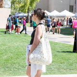 In a park, amid green scenery and people, a person in a black tank top carries a Bumkins Packable Tote Bag: Palm Daze.