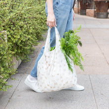 Load image into Gallery viewer, A person in jeans and white sneakers carries a Bumkins Packable Tote Bag: Palm Daze filled with greens, walking on a plant-lined sidewalk.
