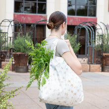Load image into Gallery viewer, A woman with a bun carries a Bumkins Packable Tote Bag: Palm Daze filled with greens outside a building with decorative metal arches.
