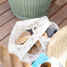 Load image into Gallery viewer, A Bumkins Packable Tote Bag: Palm Daze with flip-flops, hat, and clothes rests on a wooden surface near a green planter.
