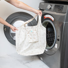 Load image into Gallery viewer, A person holds Bumkins Packable Tote Bag: Palm Daze near an open front-loading washer.
