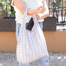 Load image into Gallery viewer, Person places a phone into Bumkins Packable Tote Bag: Lunar Phase while wearing a sleeveless sweater and jeans.
