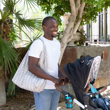 Load image into Gallery viewer, A man in a white shirt pushes a stroller with Bumkins Packable Tote Bag: Lunar Phase on his shoulder in a sunny outdoor setting with trees.
