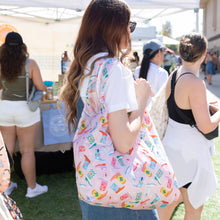 Load image into Gallery viewer, A person with long hair holds a Bumkins Packable Tote Bag: Boots at an outdoor market, among bustling crowds and vibrant vendor booths.
