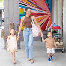 Load image into Gallery viewer, A woman walks with two kids, holding a Bumkins Packable Tote Bag: Boots, near a colorful mural. Its sunny and they are all smiling.
