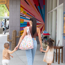 Load image into Gallery viewer, A woman with a Bumkins Packable Tote Bag: Boots and two children strolls by a mural near a cactus-shaped art piece.
