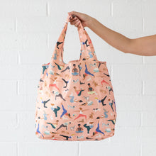 Load image into Gallery viewer, A hand holds a Bumkins Packable Tote Bag: Yoga Pals against a white brick wall.
