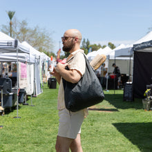 Load image into Gallery viewer, A man with a Bumkins Packable Tote Bag: Rich Black, baguettes, and iced coffee strolling through a sunny outdoor market with white tents.
