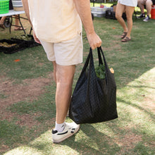 Load image into Gallery viewer, A person in shorts and sneakers holding a Bumkins Packable Tote Bag: Rich Black with greens at an outdoor market.
