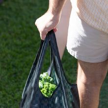 Load image into Gallery viewer, Close-up of a person in shorts holding a Bumkins Packable Tote Bag: Rich Black, with fresh broccoli visible. Grass is seen in the background.
